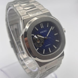 May include: A silver-toned stainless steel Seiko automatic watch with a blue horizontal striped dial. The watch features a circular bezel, a metallic link bracelet, and an open-heart aperture at the nine o'clock position displaying the movement.