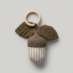 May include: A plush acorn toy featuring a textured brown cap and a light beige and white striped body. Two olive green fabric leaves with stitched vein details are attached to the top, which is connected to a smooth, round wooden ring.