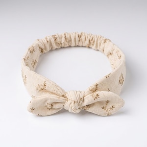Peut inclure: Un bandeau en tissu beige orné d'un petit motif floral marron sur toute la surface. L'accessoire présente un nœud noué au centre sur le devant et une bande élastique à l'arrière pour assurer un maintien sûr autour de la tête de l'utilisateur.