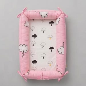 May include: A rectangular baby nest with a solid pink exterior and a white interior featuring a repeating pattern of black and yellow clouds, rain, and lightning bolts. The padded sides are secured at each corner with pink fabric ties.