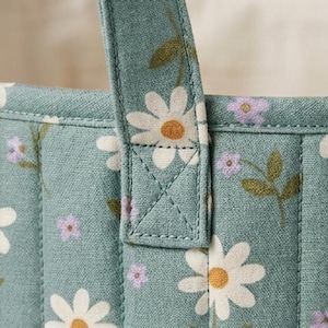 May include: A quilted fabric tote bag in a muted sage green color featuring a repeating pattern of white daisies with yellow centers and small light purple flowers. The bag has a vertical stitched handle attachment with a reinforced X-stitch.