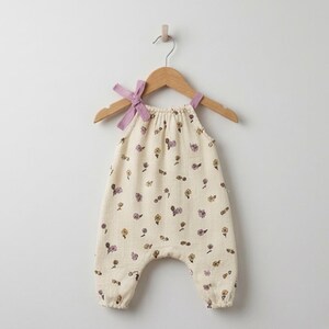 May include: A cream-coloured romper featuring a delicate floral pattern in shades of purple and yellow. The garment has a gathered neckline and thin shoulder straps that tie into bows at the top, hanging on a wooden hanger against a plain wall.