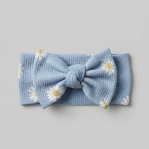Puede incluir: Una diadema de color azul claro con textura y un patrón repetitivo de margaritas blancas con centros amarillos. La banda de tela está fruncida en el centro para formar un lazo grande y prominente que se sitúa horizontalmente en el accesorio.