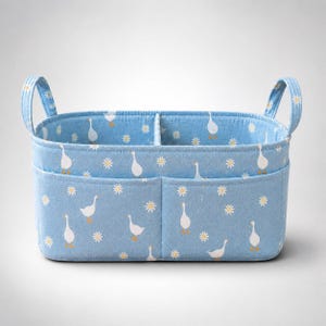 May include: A light blue fabric storage basket featuring a repeating pattern of white geese and yellow daisies. The rectangular organiser includes two side handles, a central interior divider, and two exterior pockets on the front panel.