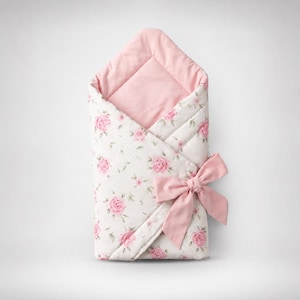 Könnte beinhalten: Ein gesteppter Baby-Wickel mit einem Muster aus rosa Rosen und grünen Blättern auf weißem Stoff. Die Innenseite ist mit einfarbigem, hellrosa Stoff gefüttert, und die Außenseite wird mit einer passenden hellrosa Stoffschleife verschlossen.