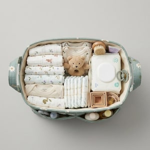 May include: An oval sage green fabric storage basket with a white daisy pattern containing baby essentials including folded muslin cloths, a beige teddy bear rattle, a pack of baby wipes, diapers, wooden stacking blocks, and two pacifiers.