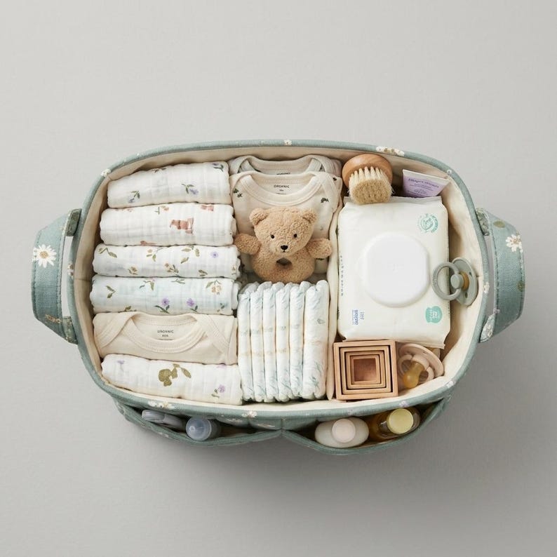 May include: An oval sage green fabric storage basket with a white daisy pattern containing baby essentials including folded muslin cloths, a beige teddy bear rattle, a pack of baby wipes, diapers, wooden stacking blocks, and two pacifiers.