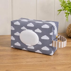 May include: A rectangular gray fabric wipe dispenser case featuring a repeating white cloud pattern. The case includes a central white plastic flip-top lid for access and a beige and white striped fabric handle attached to the side of the pouch.