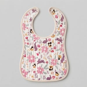 May include: A quilted baby bib featuring a repeating pattern of pink flowers, purple rabbits, and green leaves on a white background. The bib has a rounded neckline and is finished with a light beige fabric trim along the entire outer edge.