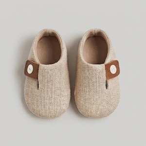 May include: A pair of beige slip-on shoes featuring a textured herringbone fabric pattern. Each shoe has a brown leather-like side strap secured with a single white button. The interior lining is a light tan colour, and the shoes have a rounded toe.