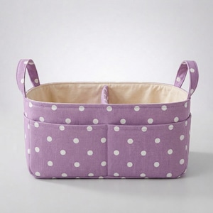 Puede incluir: Una cesta de almacenamiento rectangular de color lavanda con un patrón de lunares blancos. La cesta incluye dos bolsillos exteriores en la parte frontal, dos asas verticales en los laterales y un divisor interior que separa el compartimento.