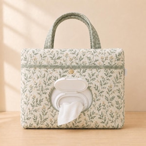 Puede incluir: Un estuche rectangular de tela acolchada para toallitas con un estampado floral botánico en verde salvia y crema. El estuche incluye dos asas superiores, una tapa abatible de plástico blanco y una toallita blanca que sobresale.