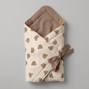 May include: A cream-colored swaddle blanket featuring a repeating pattern of small brown teddy bear faces. The blanket has a solid brown interior lining and is secured on the side with a matching brown ribbon tied into a decorative bow.