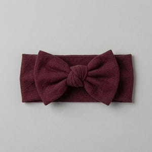 May include: A deep burgundy headband featuring a large, centered fabric bow with a knotted detail. The accessory is crafted from a soft, textured material with a wide band design that maintains a consistent color and finish throughout the piece.
