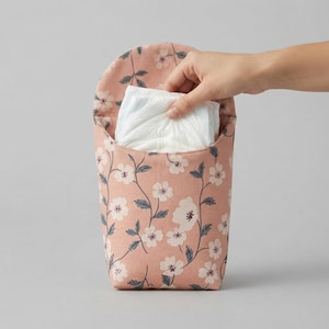 May include: A dusty pink fabric pouch featuring a repeating white floral pattern with dark green leaves. The pouch has a rounded flap and contains a folded white disposable diaper, which is partially visible at the top opening of the container.