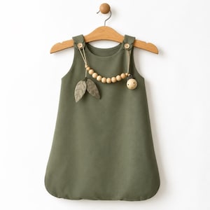 May include: An olive green sleeveless dress hanging on a wooden hanger. The dress features two small wooden buttons at the shoulders, from which a string of light-colored wooden beads is suspended, accented by two fabric leaves and a wooden sphere.