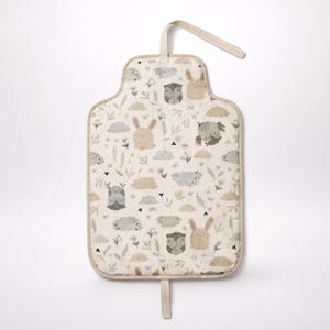 Puede incluir: Un cambiador portátil rectangular con fondo beige que presenta un patrón repetitivo de animales del bosque en gris y beige, incluyendo conejos, búhos y erizos, intercalados con pequeñas nubes, hojas y diminutos corazones negros.
