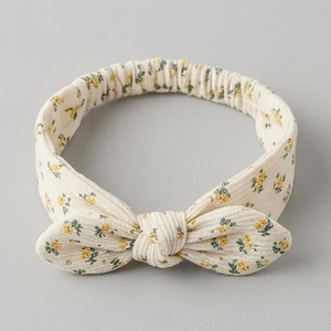 Puede incluir: Una diadema de pana de color crema con un pequeño estampado floral amarillo y verde. La diadema presenta un detalle de lazo anudado en la parte delantera y una banda elástica en la parte posterior para asegurar un ajuste firme.