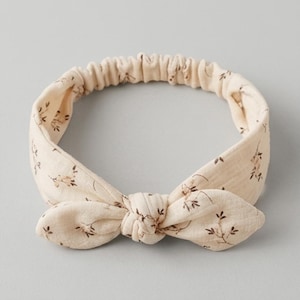 Könnte beinhalten: Ein cremefarbenes Stirnband mit einem kleinen braunen Blumenmuster. Das Stirnband verfügt über eine geknotete Schleife auf der Vorderseite und ein elastisches Band auf der Rückseite für einen sicheren Sitz. Der Stoff hat eine weiche, strukturierte Optik.
