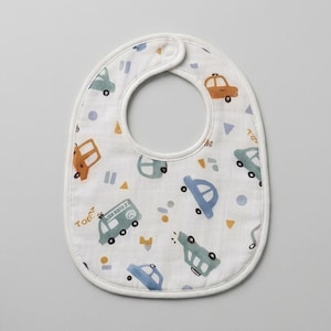 Puede incluir: Un babero blanco para bebé con un patrón repetitivo de coches pequeños, furgonetas y formas geométricas en tonos apagados de azul, verde y naranja. El babero tiene una forma redondeada con un borde de tela blanca y un cierre de presión.