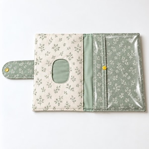 Puede incluir: Un estuche rectangular de tela para toallitas con un exterior de color crema con un patrón de hojas botánicas verdes y un interior verde a juego. El estuche incluye una abertura ovalada central, una solapa lateral con un broche amarillo y un bolsillo transparente.
