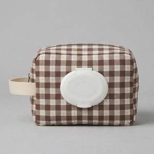 May include: A rectangular fabric pouch featuring a brown and cream gingham check pattern. The front of the pouch has a white plastic flip-top dispenser lid centered on the surface. A beige woven fabric strap is attached to the left side panel.