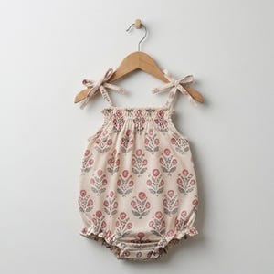 Puede incluir: Un pelele de color crema con un patrón floral repetitivo en tonos rojo apagado y gris. La prenda cuenta con un corpiño fruncido, tirantes finos que se atan con lazos y aberturas para las piernas con volantes, colgado en una percha de madera.