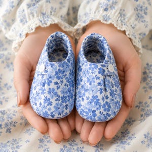 Op de afbeelding: Een paar babyschoentjes met een blauw-wit bloemenpatroon. De schoenen hebben een ronde neus, een instapmodel en een klein wit knoopdetail aan de zijkant van elke schoen. De stof is bedekt met een dichte print van kleine blauwe bloemetjes.