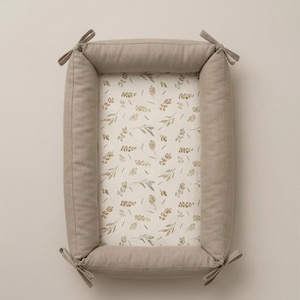 May include: A rectangular baby nest with solid beige padded sides and fabric ties at each corner. The interior base features an off-white fabric printed with a repeating pattern of small, muted green and brown botanical leaf and sprig illustrations.