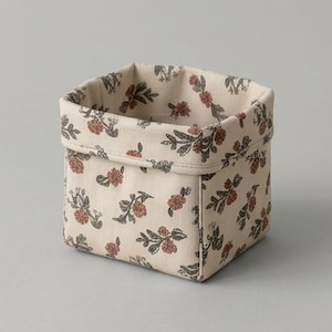 May include: A square fabric storage bin featuring a cream-colored base with a repeating pattern of small, muted orange flowers and dark green leaves. The container has a folded top edge and a structured, boxy shape with visible stitched seams.
