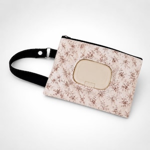 May include: A quilted rectangular pouch featuring a light pink fabric with a brown floral pattern. The pouch includes a black zipper, a black wrist strap with a silver metal ring, and a central beige plastic flip-top opening for dispensing wipes.