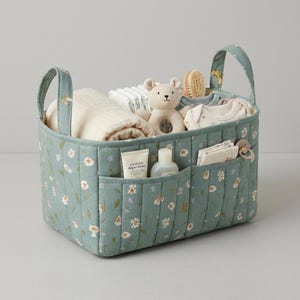 May include: A sage green quilted fabric storage basket with two handles and a white floral pattern. The basket contains a beige plush bear toy, a folded cream blanket, a white baby bodysuit, a small bottle, and a pacifier with a clip.