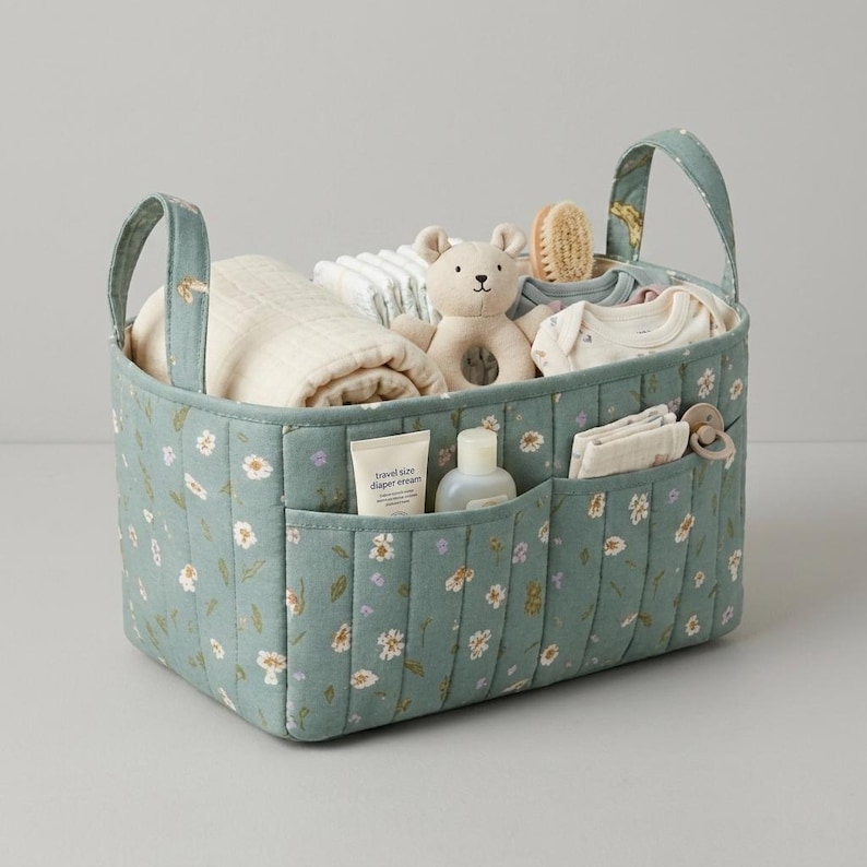 May include: A sage green quilted fabric storage basket with two handles and a white floral pattern. The basket contains a beige plush bear toy, a folded cream blanket, a white baby bodysuit, a small bottle, and a pacifier with a clip.