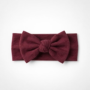 May include: A deep burgundy headband featuring a large, centred fabric bow. The accessory is crafted from a soft, textured knit material with a consistent dark red colour throughout, showcasing a knotted centre detail on the prominent bow.