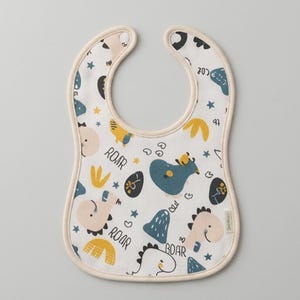 May include: A white baby bib featuring a repeating pattern of cartoon dinosaurs in shades of blue, yellow, and light pink. The design includes small stars, abstract shapes, and the word ROAR printed in black text throughout the fabric surface.