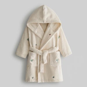 May include: A cream-colored hooded bathrobe made of soft terry cloth fabric. The robe features long sleeves, a matching tie belt at the waist, and two front patch pockets. Small, colorful embroidered icons are scattered across the fabric.