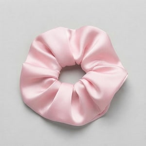May include: A light pink hair scrunchie with a smooth, satin-like finish. The fabric is gathered into soft, voluminous folds around a circular elastic band, creating a rounded, pleated appearance with a consistent pastel colour throughout.