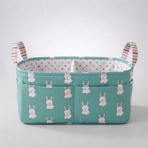 May include: A rectangular fabric storage caddy in teal featuring a repeating white bunny pattern. The caddy has two exterior pockets, a divided interior lined with white fabric featuring colourful polka dots, and two striped fabric handles.