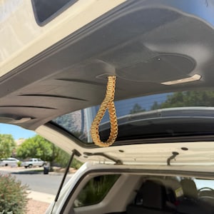 May include: A braided loop made of tan and gold cord hangs from the interior of a car's open boot. The loop is suspended from the ceiling of the boot, which is open to reveal a sunny outdoor scene.
