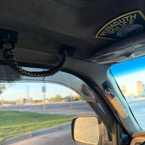 May include: Interior view of a vehicle, featuring a paracord grab handle and a Plymouth Police patch. The handle is attached to the vehicle's ceiling, with a view of the road and sky visible through the window.