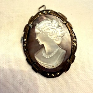 Op de afbeelding: Ovale cameohanger met een vrouwenprofiel, wit gesneden tegen een donkere achtergrond. De camee is gezet in een decoratief, sierlijk frame met een donkere metalen afwerking en kleine, sprankelende accenten. Een klassiek sieraad.