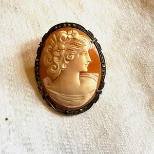 Peut inclure: Broche camée ovale représentant le profil d'une femme, avec des détails fins dans ses cheveux et ses vêtements. Le camée est monté sur un fond orange, encadré d'une bordure en métal foncé avec des accents décoratifs. Un bijou classique.