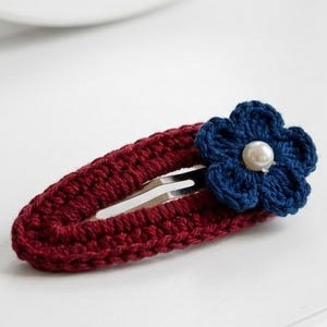 May include: A handmade crochet hair clip. The clip is a deep red color, with a navy blue flower and a pearl bead center. The clip is resting on a white surface.