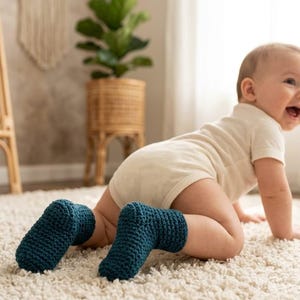 May include: A baby wearing teal blue crochet booties crawls on a cream-colored rug. The baby is wearing a white onesie. A woven basket with a green plant is in the background.