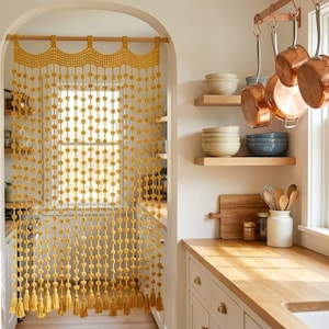 May include: A mustard yellow macrame doorway curtain featuring a scalloped top edge and multiple vertical strands adorned with small knotted spheres, ending in a series of tassels hanging from a wooden rod within an arched kitchen doorway.