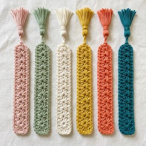 May include: Six rectangular crochet bookmarks arranged vertically, each featuring a repeating star-like stitch pattern and a matching tassel at the top. The colours from left to right are light pink, sage green, cream, mustard yellow, coral, and teal.