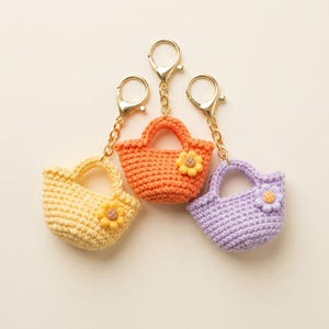 Puede incluir: Tres llaveros de mini bolsos de ganchillo en amarillo, naranja y morado claro. Cada bolso presenta una pequeña flor de ganchillo a juego en la parte delantera y está unido a un cierre de mosquetón y una cadena de metal en tono dorado.