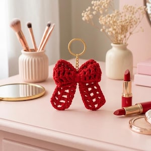 May include: A red crocheted bow keychain with a gold-colored ring. The bow is the focal point, set against a pink surface with makeup and a mirror in the background. The bow is approximately 4 inches wide.
