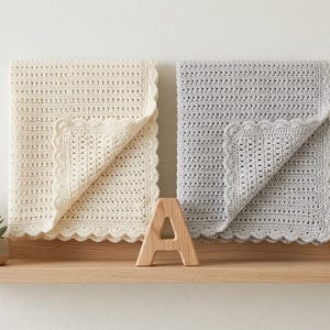 May include: Two crocheted baby blankets, one cream and one gray, displayed on a wooden shelf with decorative plants. The blankets have a scalloped edge and a textured pattern. A wooden letter "A" is placed between the blankets.