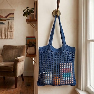 Pode incluir: Uma bolsa de crochê azul profundo com um design de malha aberta e duas alças resistentes. A bolsa está pendurada em um gancho de parede e contém uma caneca branca estampada, uma fruta laranja e um livro retangular com lombada listrada.